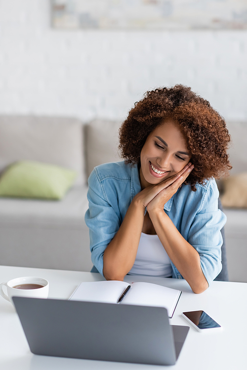 curly african american woman smiling while looking at laptop near smartphone on desk