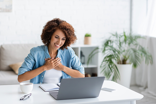 curly african american woman holding hands on chest and looking at laptop during video call