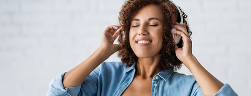 curly african american woman smiling while listening music in wireless headphones, banner