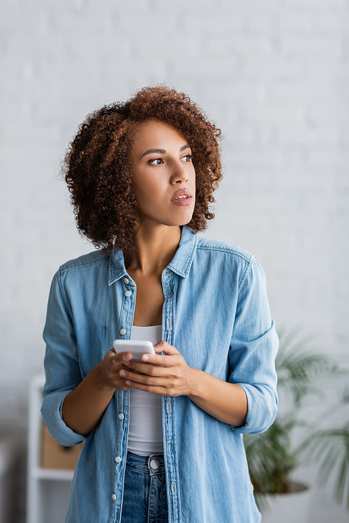 curly african american woman holding smartphone and looking away at home