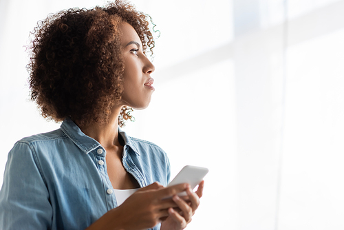 curly african american woman holding mobile phone and looking away at home