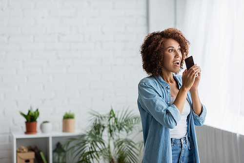 excited african american woman holding smartphone with blank screen and looking away