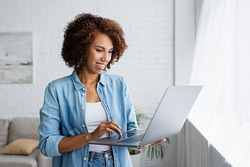 happy african american woman with curly hair holding laptop while working from home