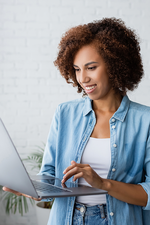 cheerful african american woman with curly hair holding laptop while working from home