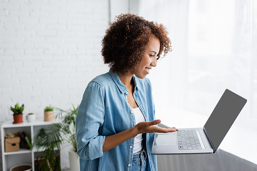 side view of smiling african american woman with curly hair holding laptop while working from home