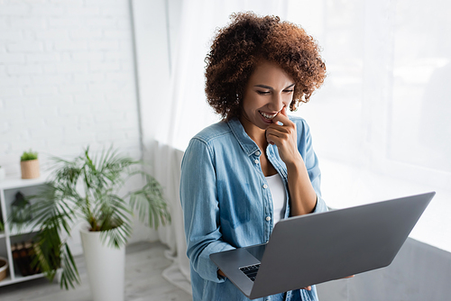 positive african american woman with curly hair holding laptop while working from home