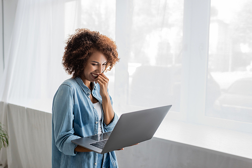 smiling african american woman with curly hair holding laptop while working from home