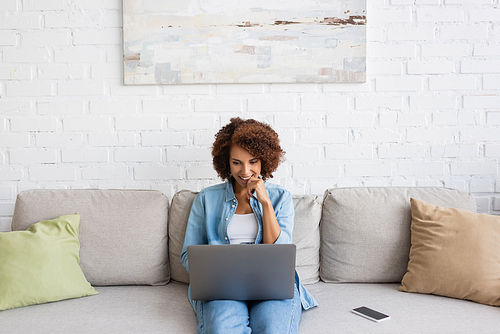 happy african american woman using laptop while sitting on couch and working from home