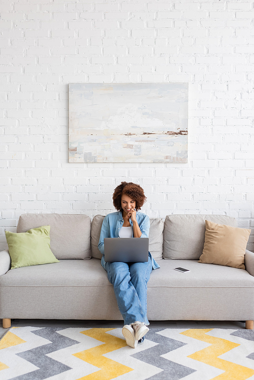 cheerful african american woman using laptop while sitting on couch and working from home