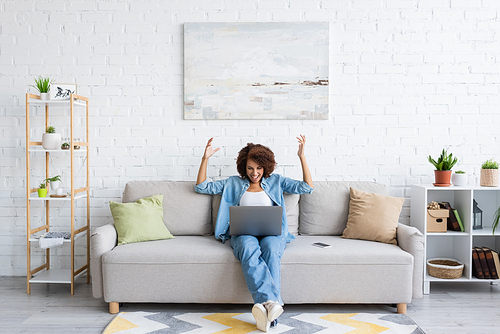 excited african american woman using laptop while sitting on couch and working from home