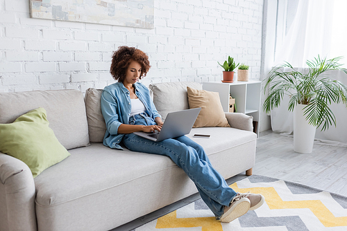 curly african american freelancer using laptop while sitting on couch and working from home