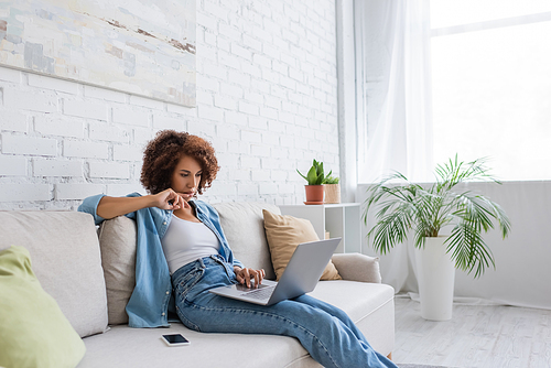 pensive african american woman using laptop while sitting on couch and working from home