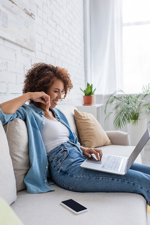 cheerful african american woman using laptop while sitting on sofa and working from home