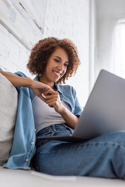 low angle view of happy african american woman using laptop while sitting on couch