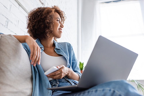 low angle view of happy african american woman holding smartphone near laptop while sitting on couch