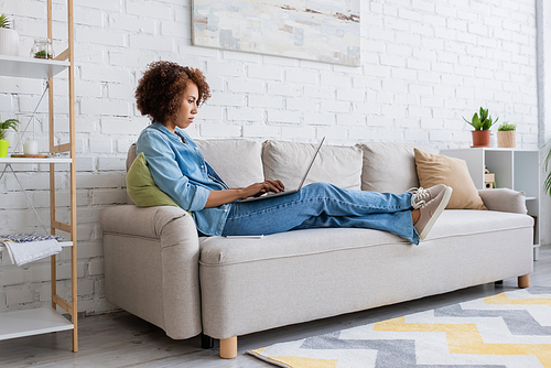 focused african american woman using laptop while sitting on couch and working from home