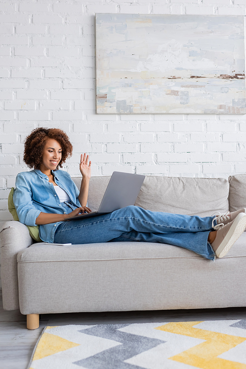 happy african american woman waving hand during video cat on laptop while sitting on couch