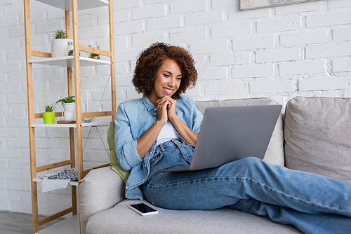 happy african american woman sitting with clenched hands and looking at laptop near smartphone on couch