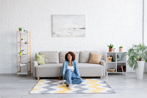 cheerful african american woman sitting on rug with pattern near modern sofa in living room