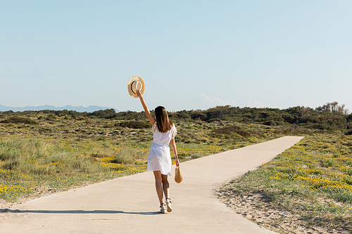 Back view of brunette woman holding sun hat and handbag while walking on beach