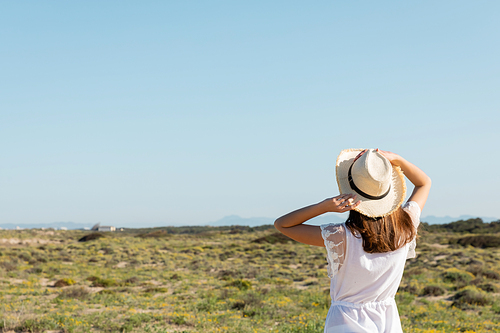 Back view of young woman holding straw hat near grass on beach at background