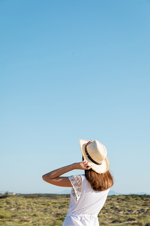 Back view of brunette woman in white dress and sun hat standing on blurred beach