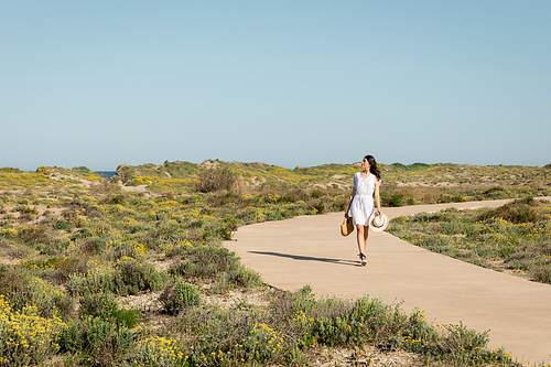 Side view of pretty woman holding handbag and straw hat while walking on pathway on beach