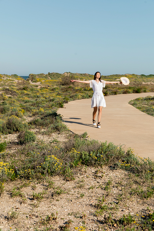 Woman in dress closing eyes and holding straw hat on pathway on beach