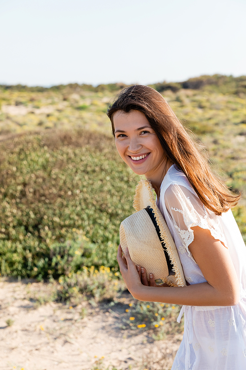 Positive woman in summer dress holding straw hat on blurred beach