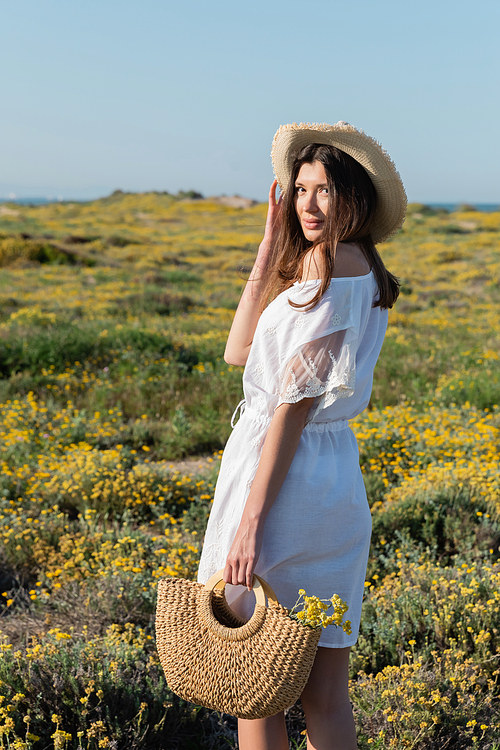 Woman in dress and sun hat holding handbag with flowers and looking at camera on beach