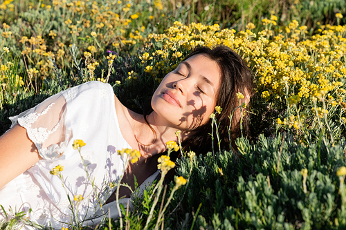 Brunette woman in white dress lying near flowers and grass on meadow