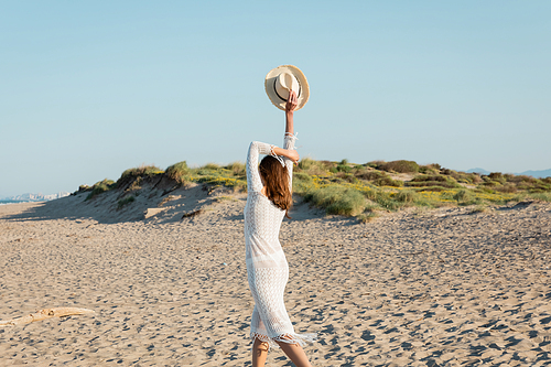 Back view of woman in knitted dress holding sun hat on sandy beach