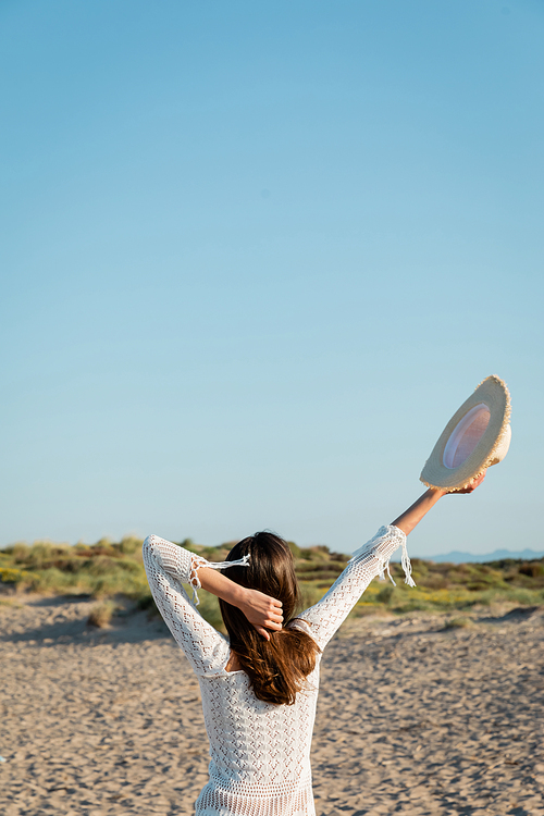 Back view of brunette woman in knitted summer dress holding sun hat and touching hair on beach