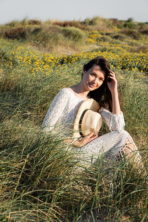 Brunette woman in summer dress holding straw hat while sitting on grass on beach