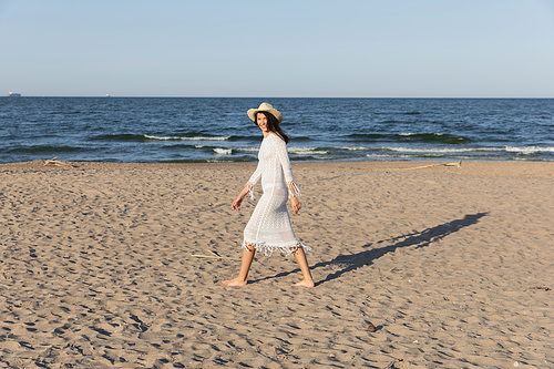 Happy woman in dress and sun hat looking at camera while walking on beach near sea