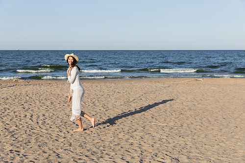 Positive woman in straw hat and summer dress walking near sea on beach