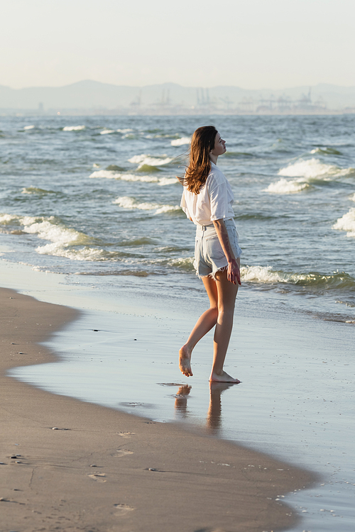 Side view of young woman in shorts standing on wet sand near sea