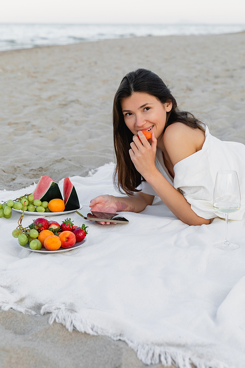 Smiling woman holding apricot and smartphone near wine and fruits on beach