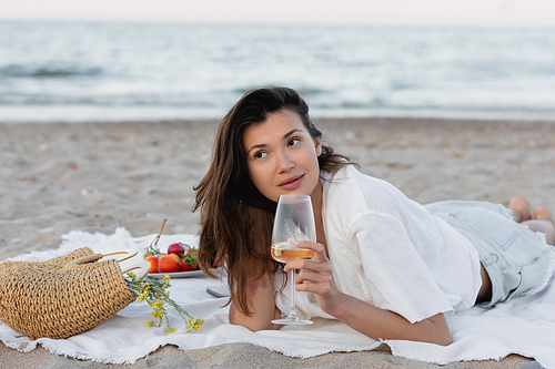 Brunette woman holding glass of wine near handbag and fruits on blanket on beach