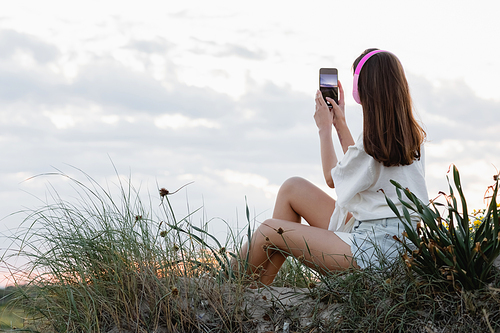 Back view of woman in headphones taking photo on smartphone on beach in evening