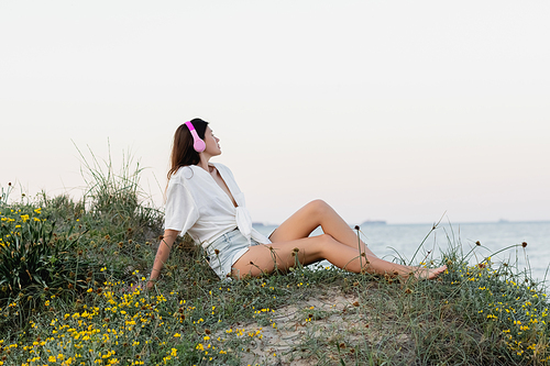 Side view of woman in headphones and shirt sitting on beach in evening