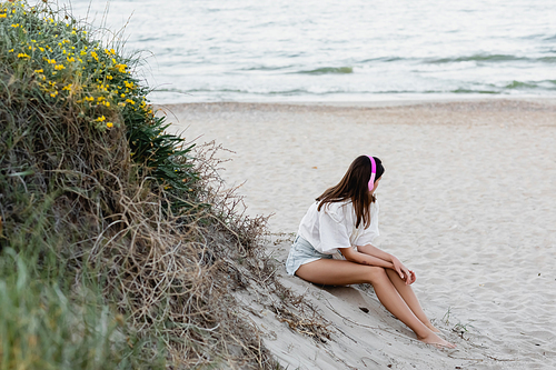 Young woman in headphones sitting on sand near sea