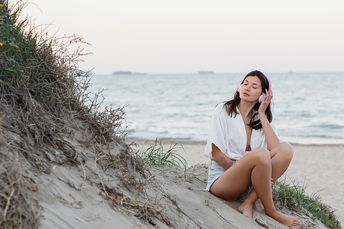 Woman in headphones closing eyes while sitting on beach in evening