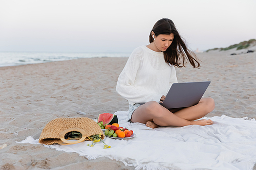 Freelancer in earphone using laptop near fruits and handbag on beach in evening