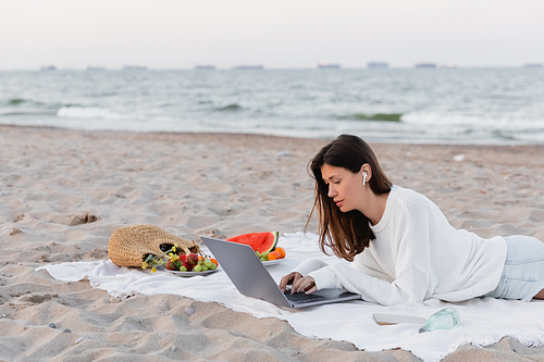 Woman in earphone using laptop near smartphone and fruits on blanket on beach