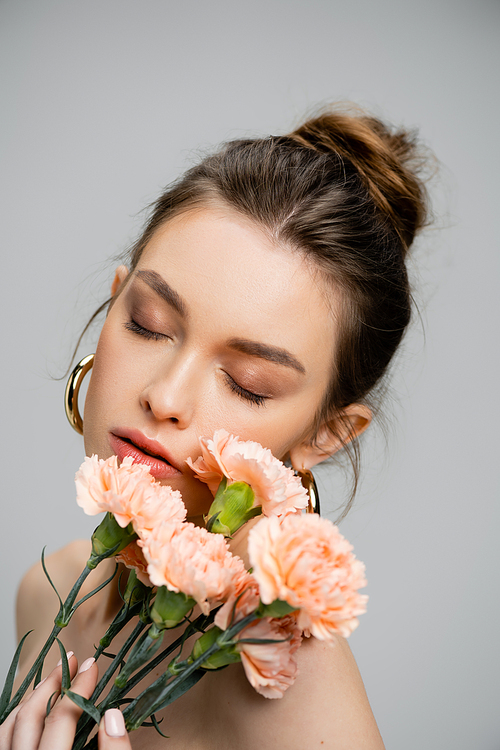 portrait of young sensual woman posing with closed eyes near bouquet of peach carnations isolated on grey