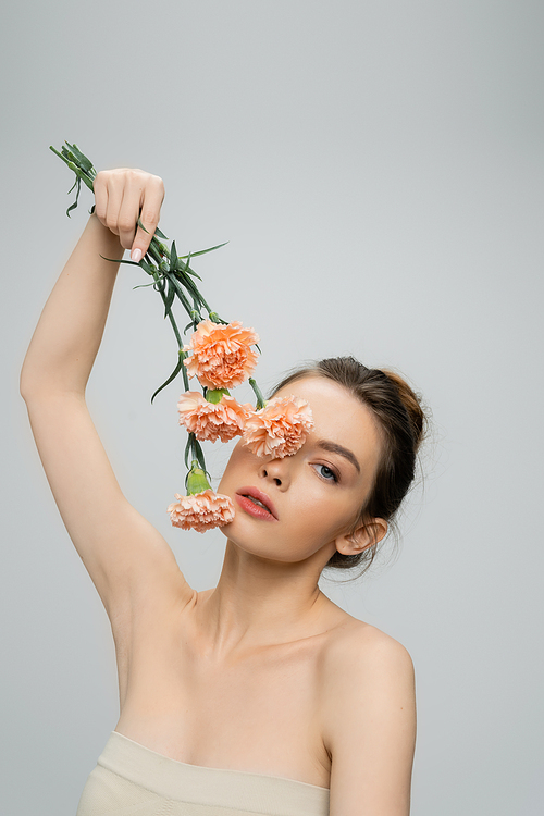 young woman with naked shoulders obscuring face with pink carnations isolated on grey