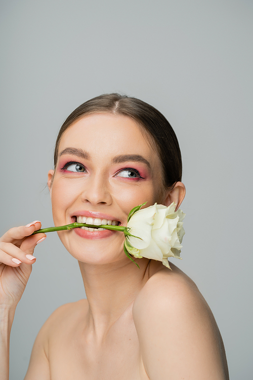 smiling woman with naked shoulders and makeup posing with white rose in teeth isolated on grey