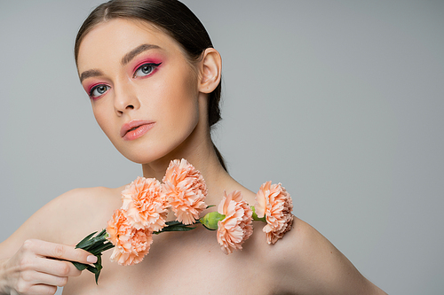 young woman with makeup and bare shoulders posing with fresh flowers and looking at camera isolated on grey