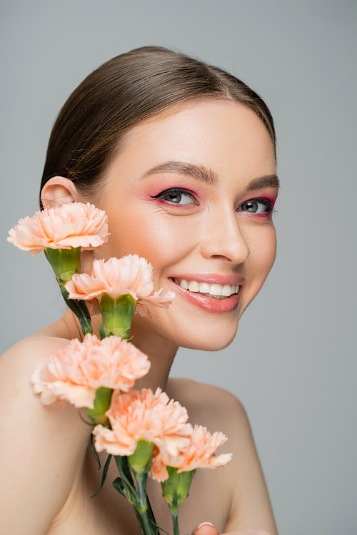 happy young woman with makeup and perfect skin smiling at camera near peach carnations isolated on grey
